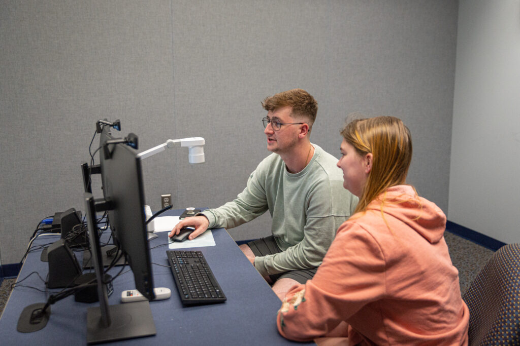 Two students sit at a desk and look at two computer screens.