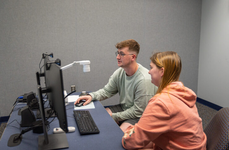 Two students sit at a desk and look at two computer screens.