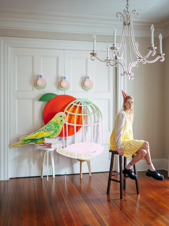 Woman wearing party hat sits on a stool with sculptures of a peach, bird, and birdcage behind her.