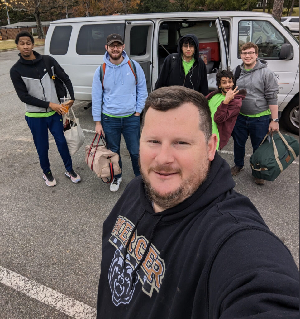 A professor takes a selfie of himself and four students in front of a van.