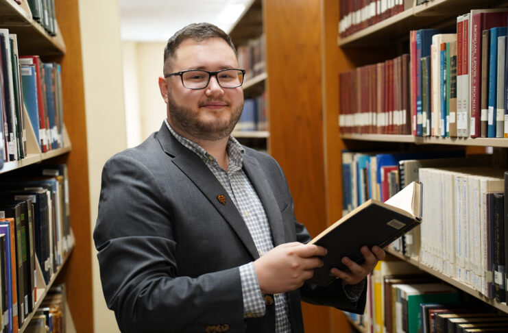 A man in a suit stands holding a book in a library