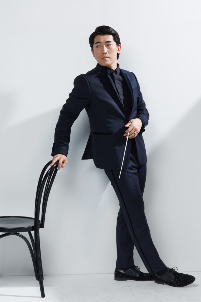 A man in a black suit touches a chair back with one hand and holds a conducting baton with the other.
