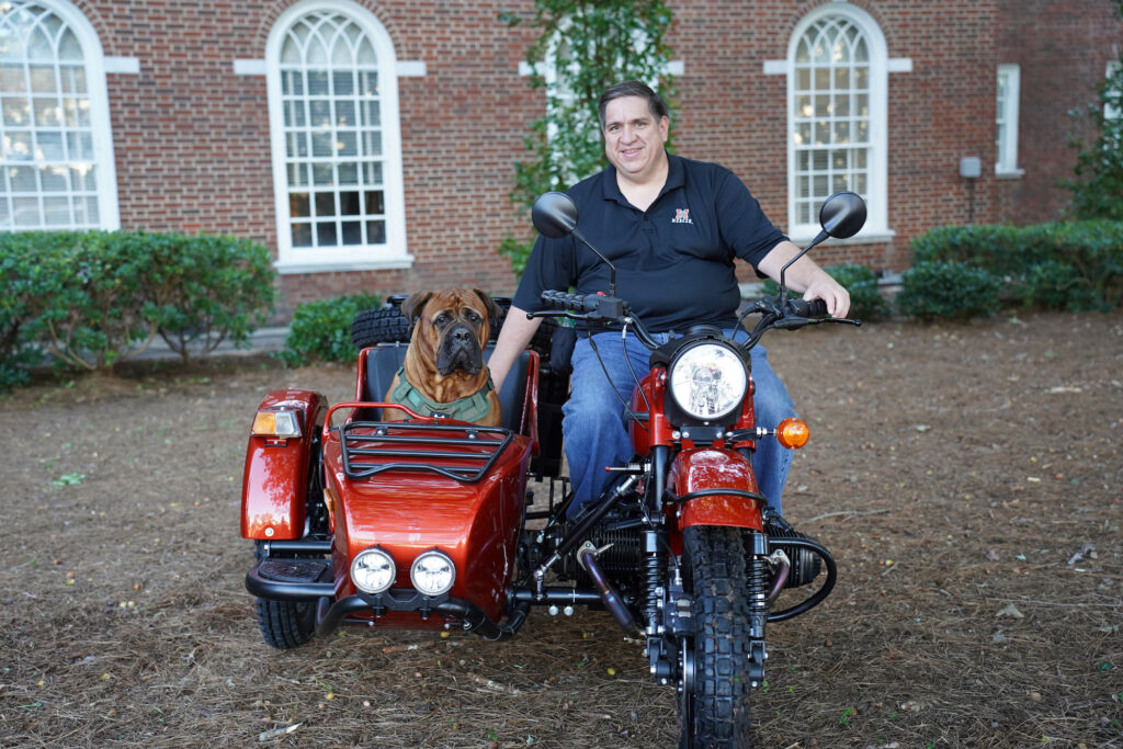 A man sits atop a motorcycle with a dog in the attached side car