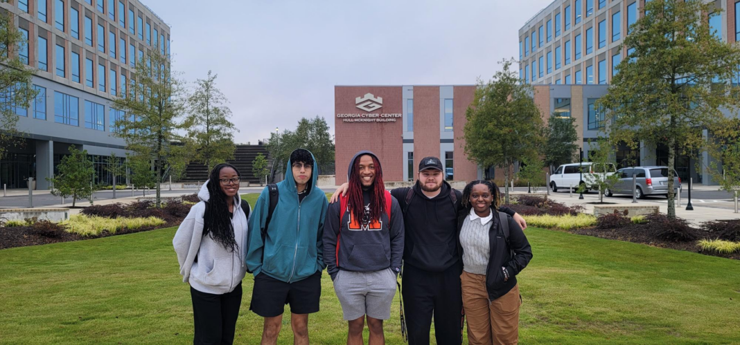 Five students stand in a row for a picture outside, in front of a brick building.