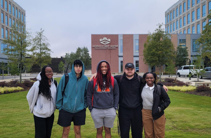 Five students stand in a row for a picture outside, in front of a brick building.