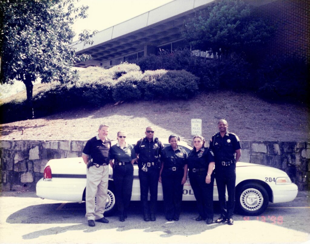 Six police officers in uniform stand in front of a police car.