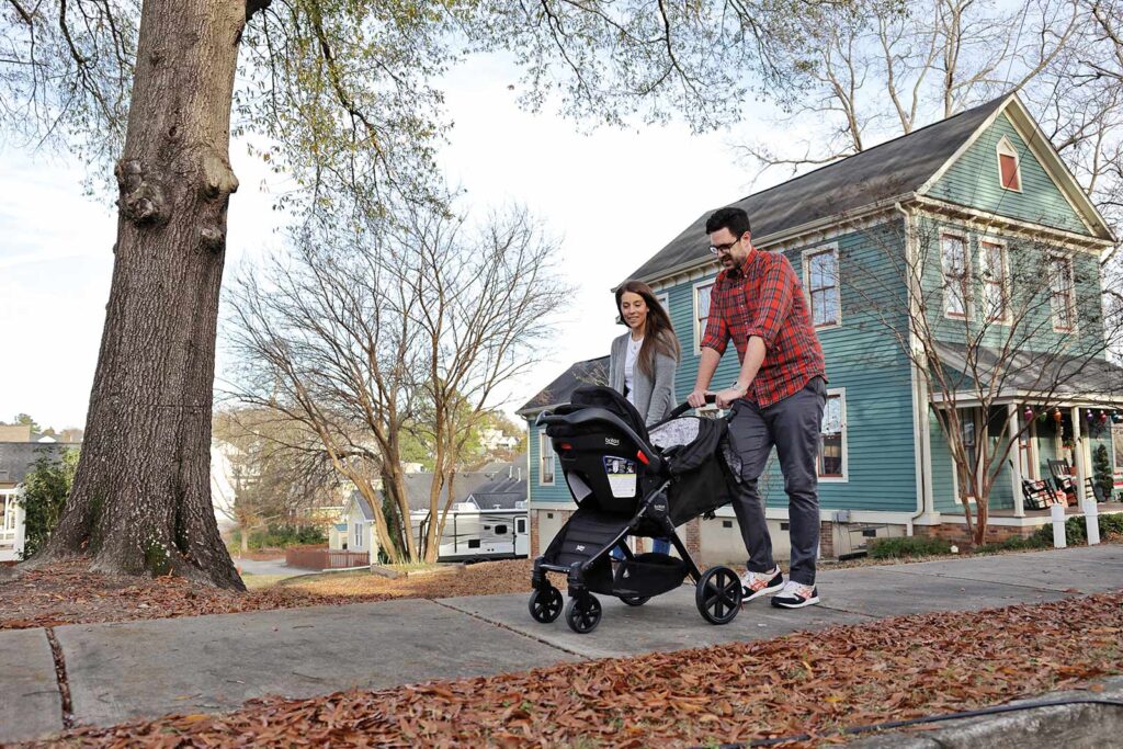 a man and woman push a baby stroller down a sidewalk with a home in the background