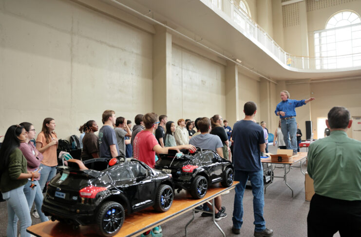 A man stands on a table to talk with a large group of students, with toy electric cars on another nearby table.