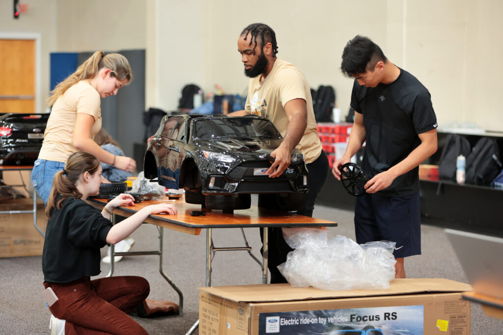 Four students surround a table and work to assemble a black toy jeep.