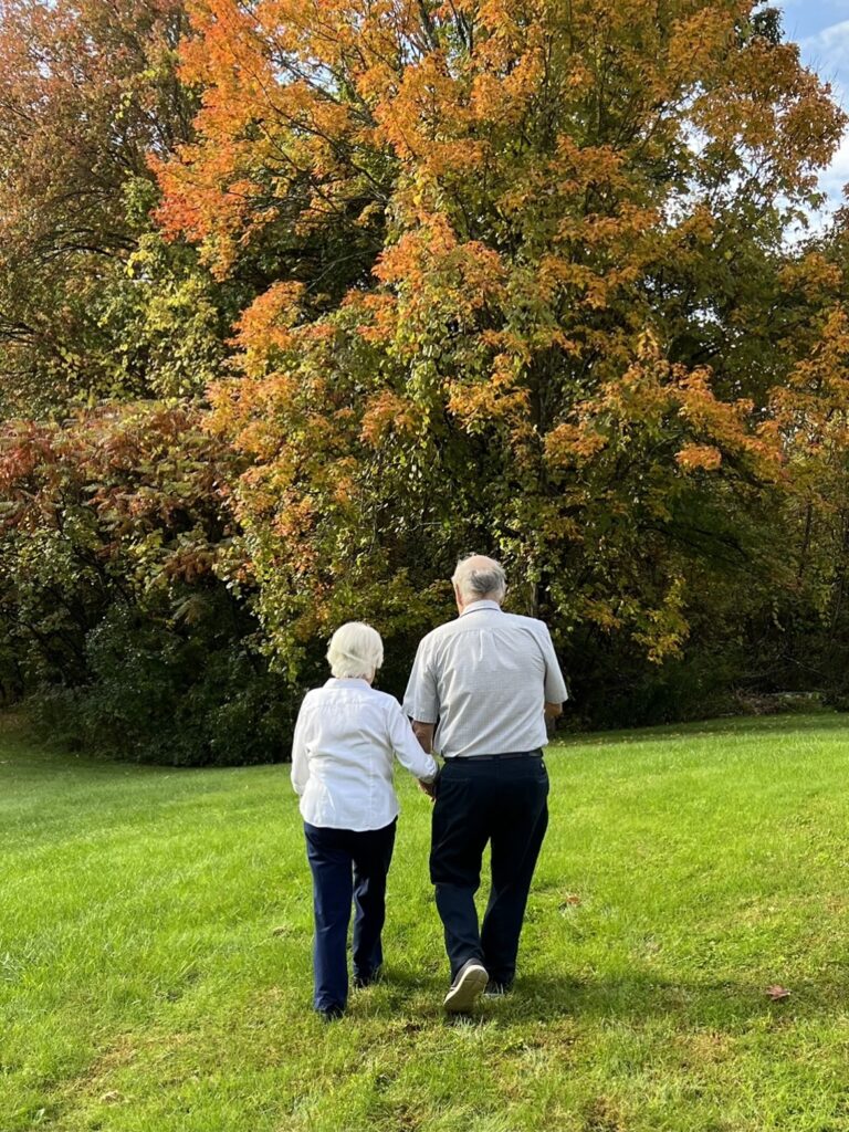 elderly couple walking in field toward trees with fall leaves