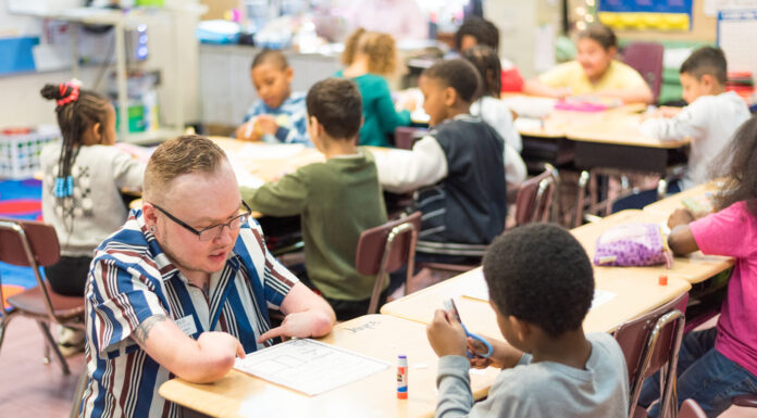 Mercer senior’s own experiences inspire him to become special education teacher a man born with shortened arms squats on the other side of a desk where a student is cutting a piece of paper