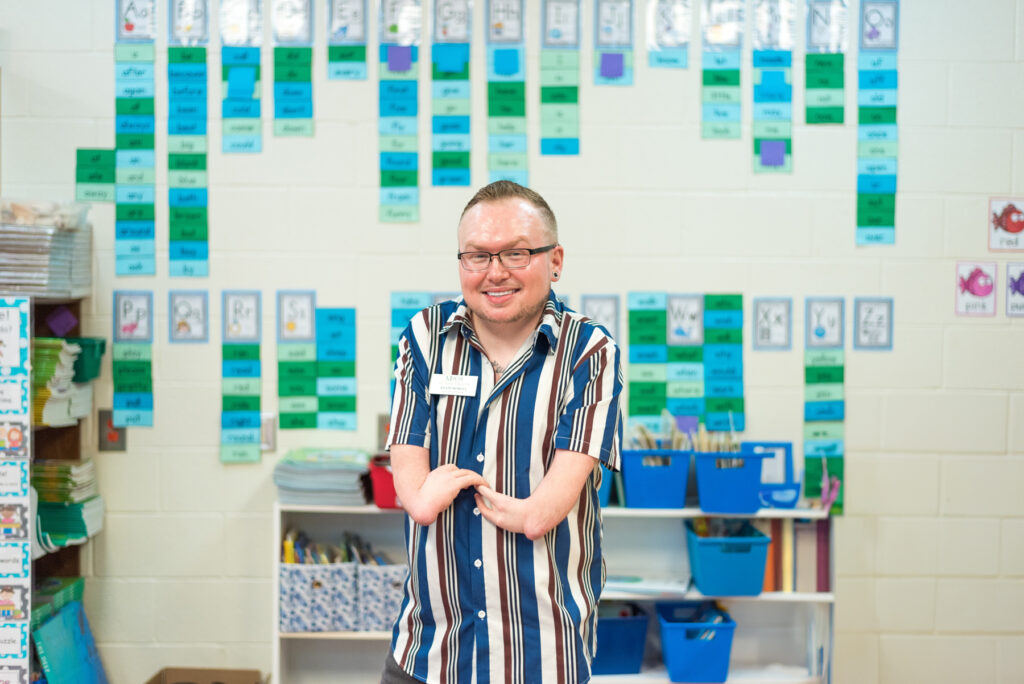 a man born with shortened arms wears a striped shirt and stands in front of a a wall with colorful notecards on it