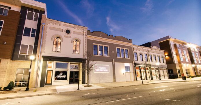 Street view of a row of modern and historic buildings with lit sidewalks and clear skies at dusk.