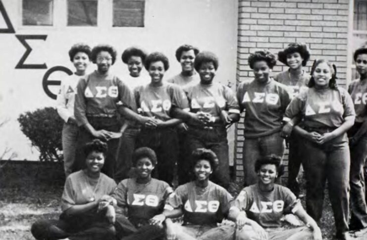 A group of sorority members in matching shirts pose for a photo outside, with linked arms and five sitting on the ground and 10 standing.
