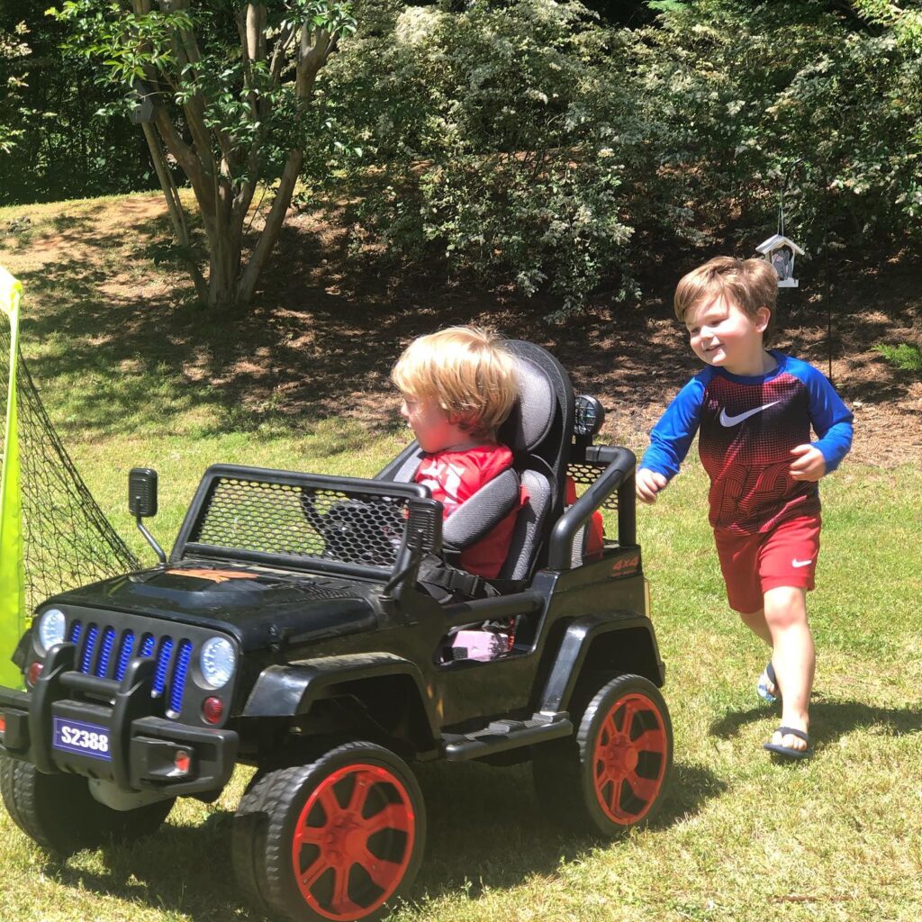 A boy drives his motorized car while his brother runs beside him.