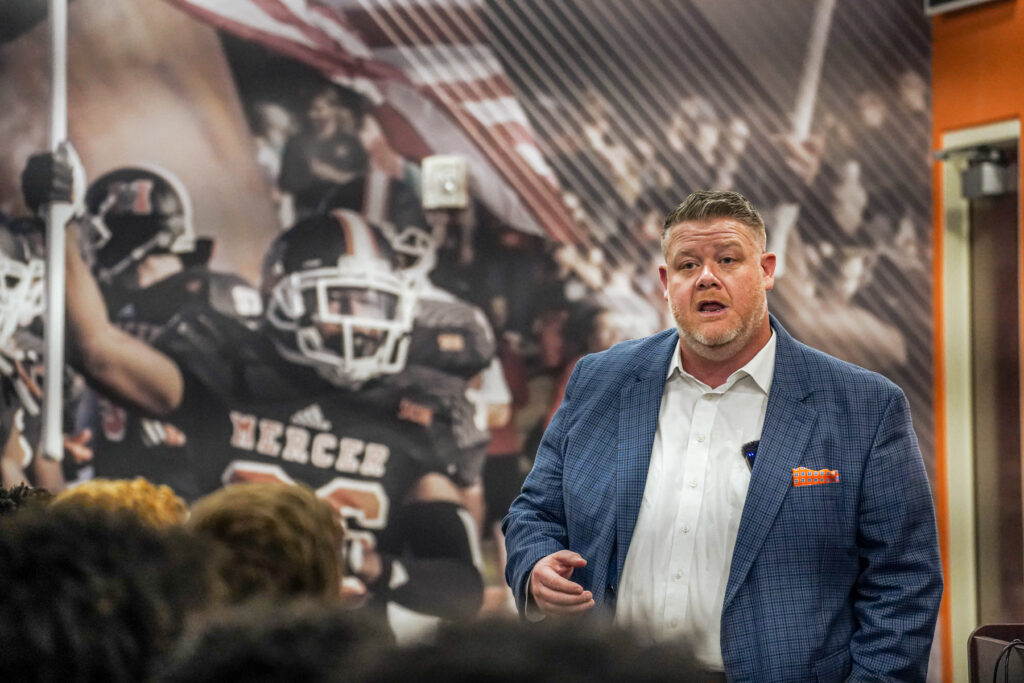 Mike Jacobs talks to a group of people. A banner behind him shows a Mercer football player waving the American flag.