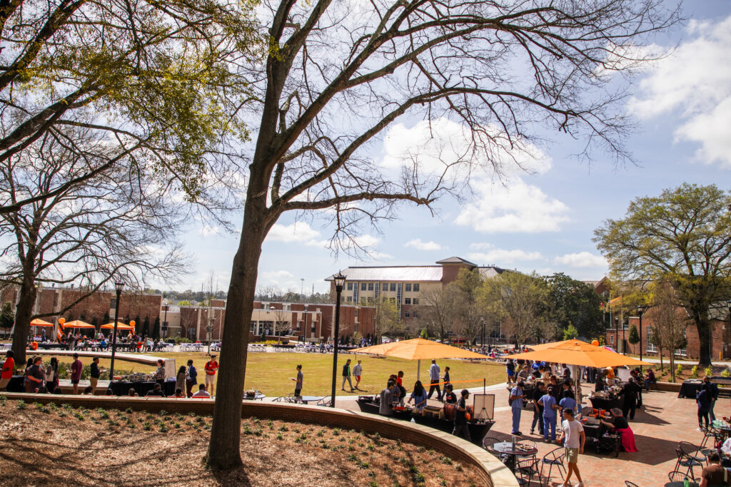 Students gather on a green space with orange table umbrellas and trees.