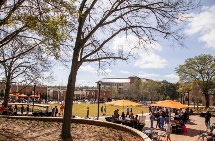 Students gather on a green space with orange table umbrellas and trees.