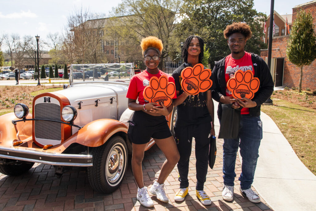 Three students hold foam Mercer Bear signs while standing in front of an orange and white car.