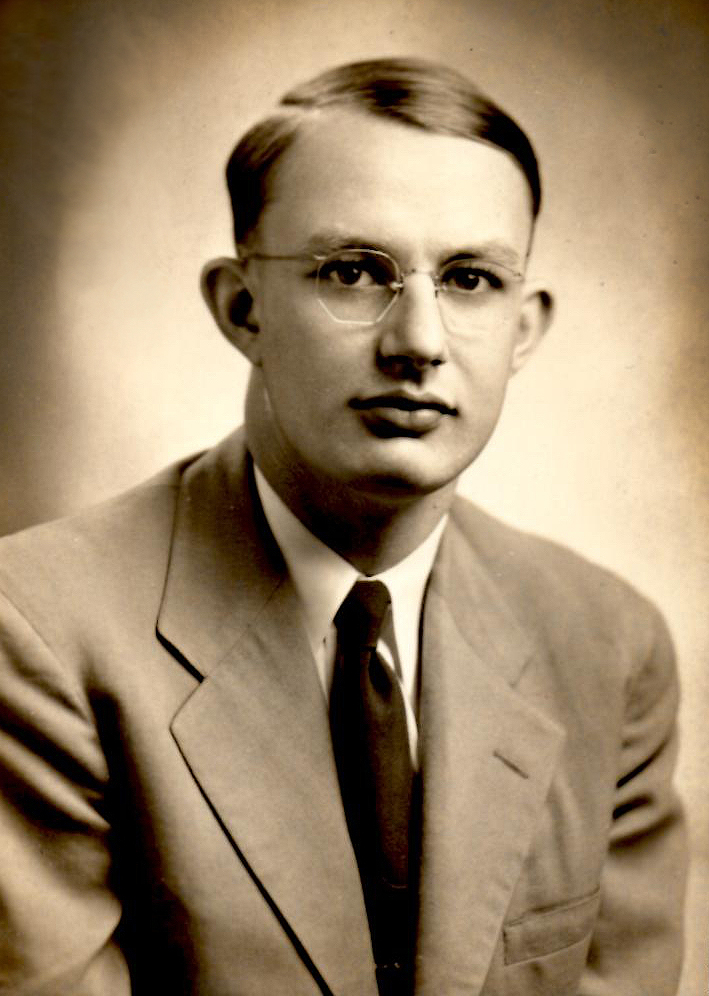 A black and white photo of a young man in glasses wearing a suit and tie.