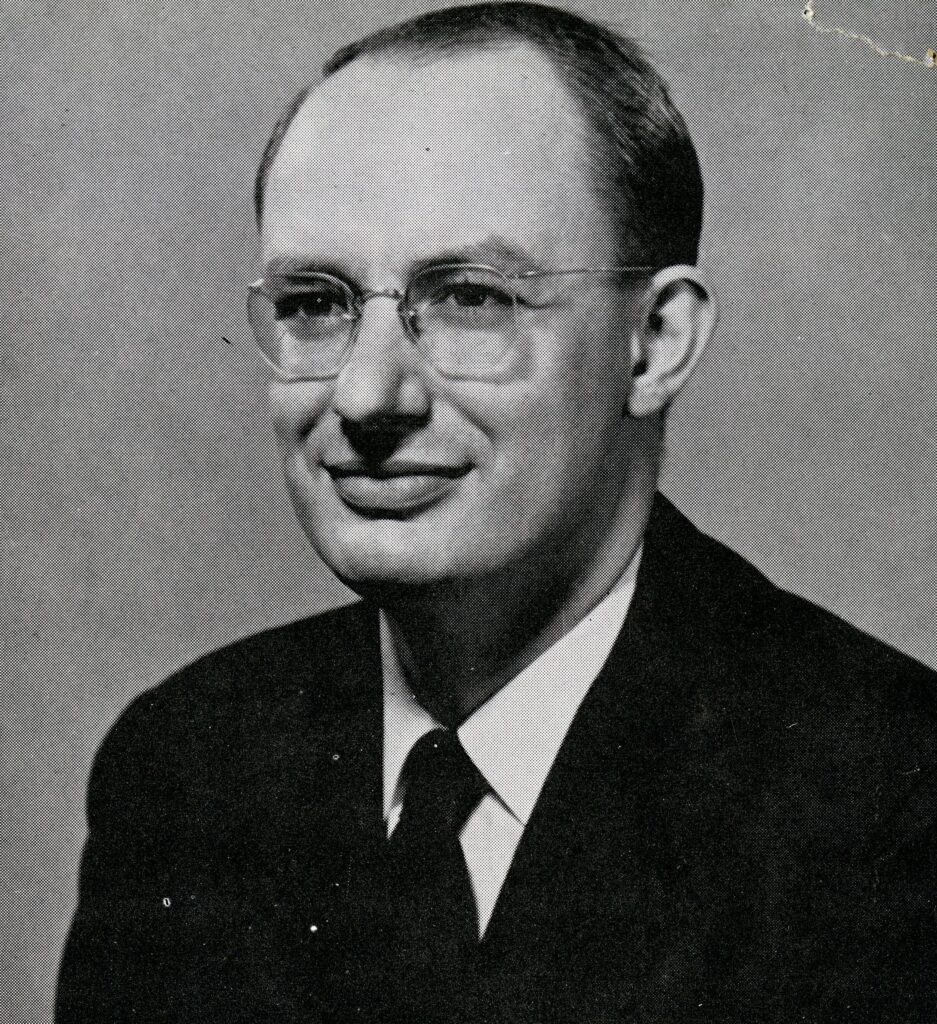 A black and white photo of a man in glasses wearing a suit and tie.