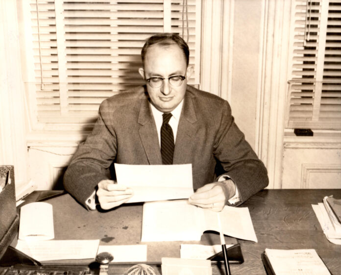 A black and white photo of a man in a suit reading papers at a desk.