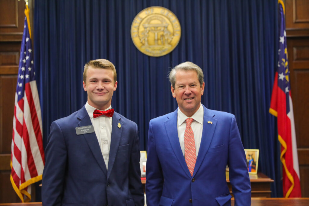 two men in suits stand next to each other in front of a large wooden desk with the Georgia state seal hanging behind them