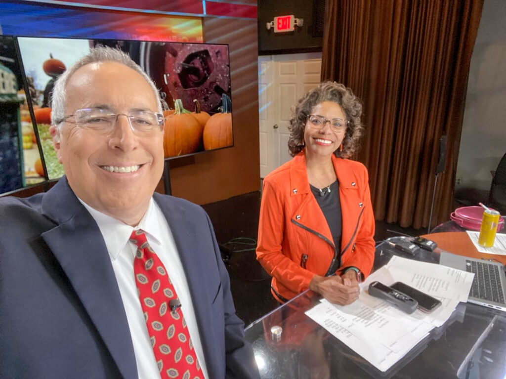 a man holds out his arm to take a selfie of himself and a woman sitting behind a news desk