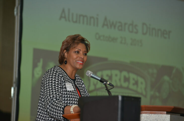 woman speaks into a microphone in front of screen with the words alumni awards dinner projected on it