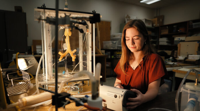 Goldwater Scholar strives for ‘excellence, innovation’ A young woman in a red shirt sits in a dark room, with nebulizer equipment around her.