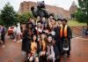 Hey 2026 graduates! We want to feature you in The Den A group of graduates in caps and gowns pose in front of a large bear statue on a brick-paved area.