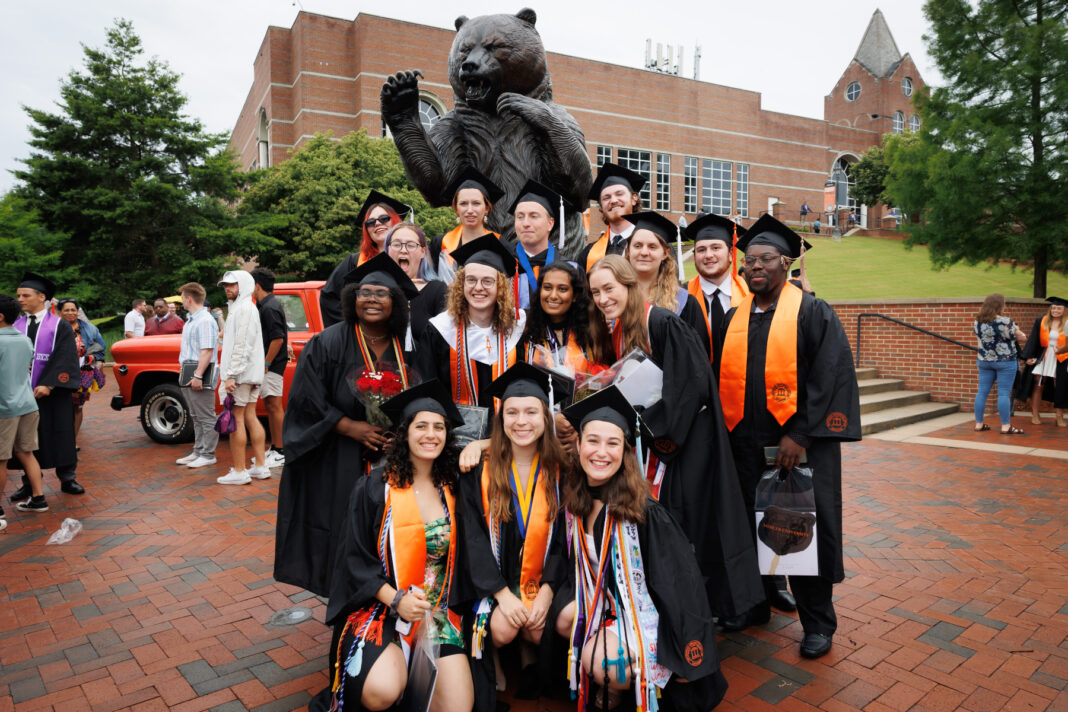 A group of graduates in caps and gowns pose in front of a large bear statue on a brick-paved area.