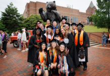 Hey 2026 graduates! We want to feature you in The Den A group of graduates in caps and gowns pose in front of a large bear statue on a brick-paved area.