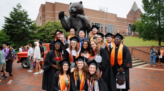 Hey 2026 graduates! We want to feature you in The Den A group of graduates in caps and gowns pose in front of a large bear statue on a brick-paved area.