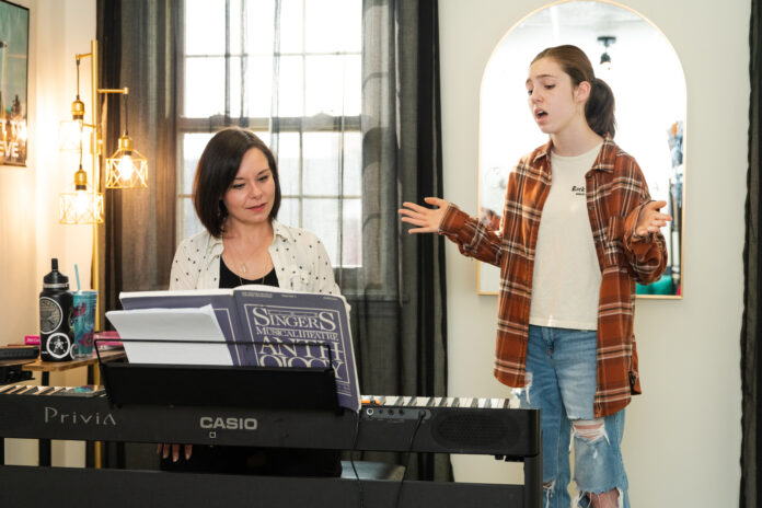 A woman plays piano while a student sings standing beside her with arms outstretched.