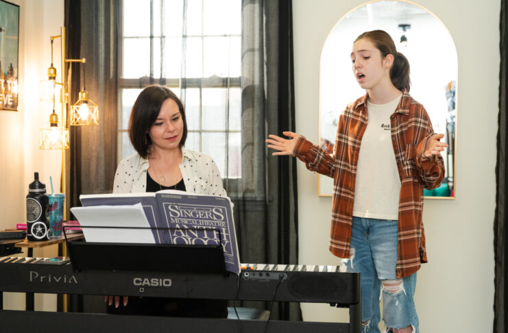 A woman plays piano while a student sings standing beside her with arms outstretched.