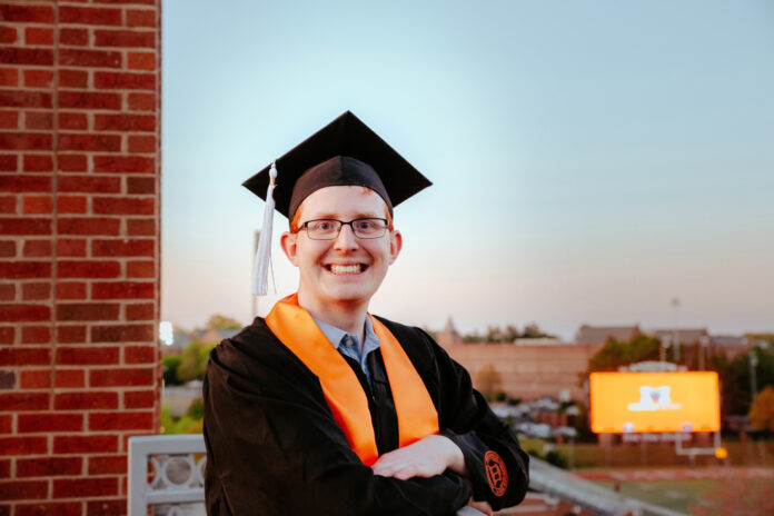 a male student wears graduation regalia with the football stadium in the background