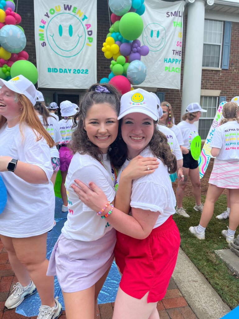 Two young women wearing colorful skirts and white shirts post for a photo, with other women and balloons in the background.