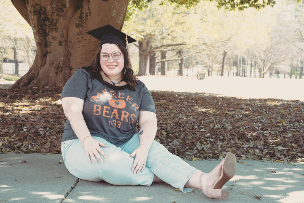 female wearing a mercer shirt and graduation cap sits in front of a tree