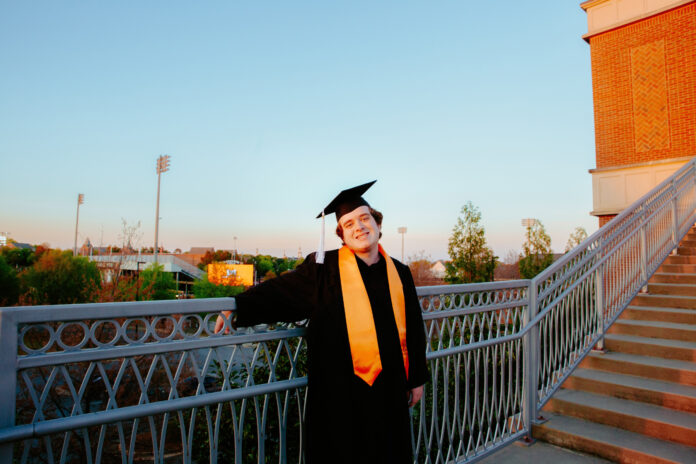 man in graduation regalia stands at a railing overlooking the mercer football stadium