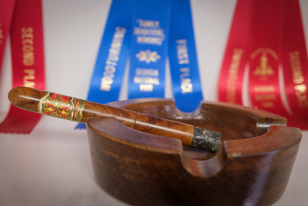 A pen made to look like a cigar sits atop an ashtray. a blue ribbon is behind it