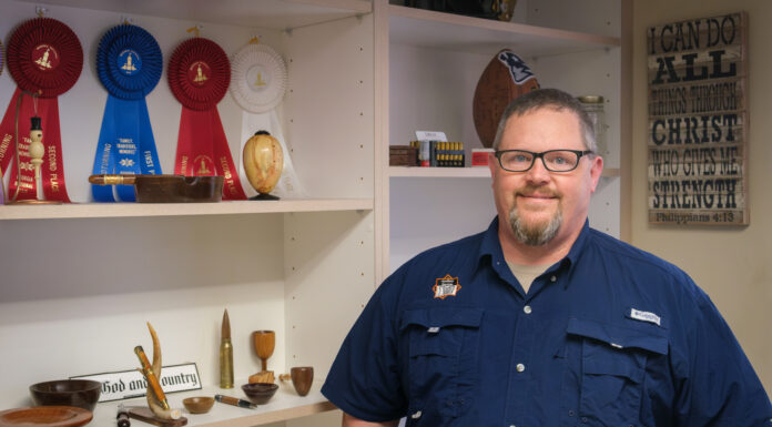 Mercer employee finds beauty, joy and God in wood turning a man stands next to a bookcase with objects he turned out of wood and prize ribbons