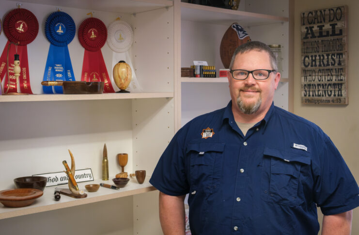 a man stands next to a bookcase with objects he turned out of wood and prize ribbons