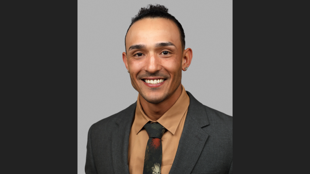 A headshot of a man in a gray suit jacket and orange shirt.