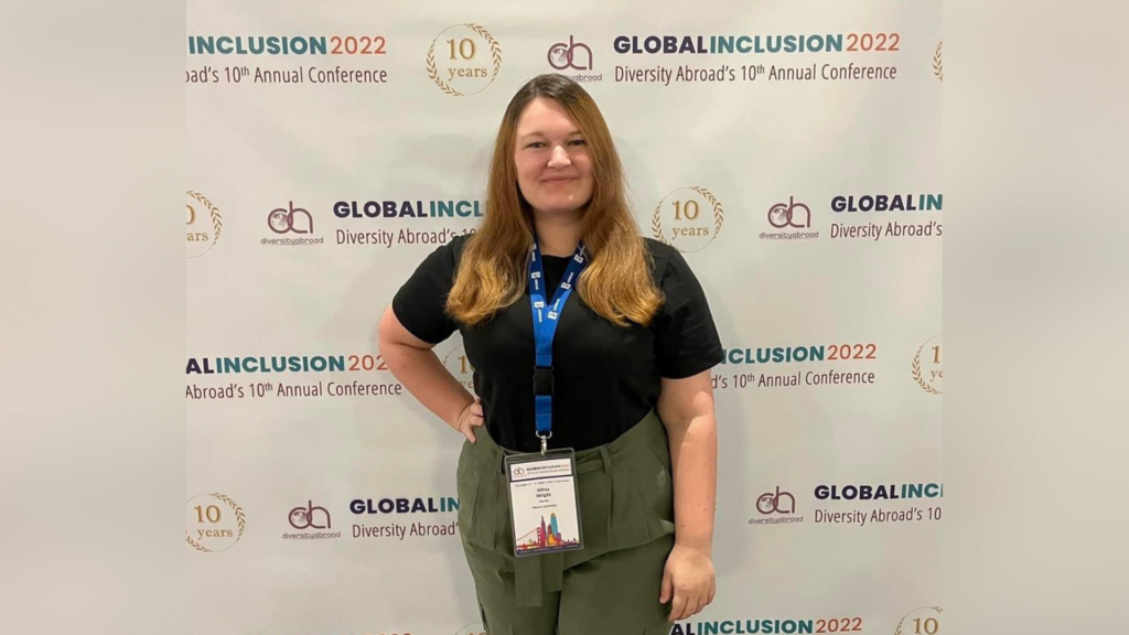 A woman with auburn hair wearing a black shirt and green pants stands in front of a backdrop that says Global Inclusion 2022 Diversity Abroad's 10th Annual Conference.