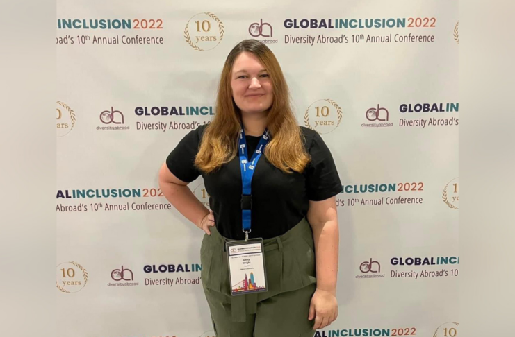 A woman with auburn hair wearing a black shirt and green pants stands in front of a backdrop that says Global Inclusion 2022 Diversity Abroad's 10th Annual Conference.