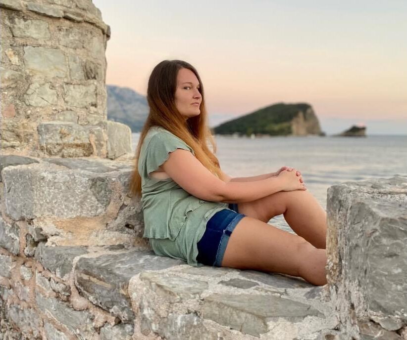 A woman wearing jean shorts and green shirt sits on a stone wall, with the ocean in the background.