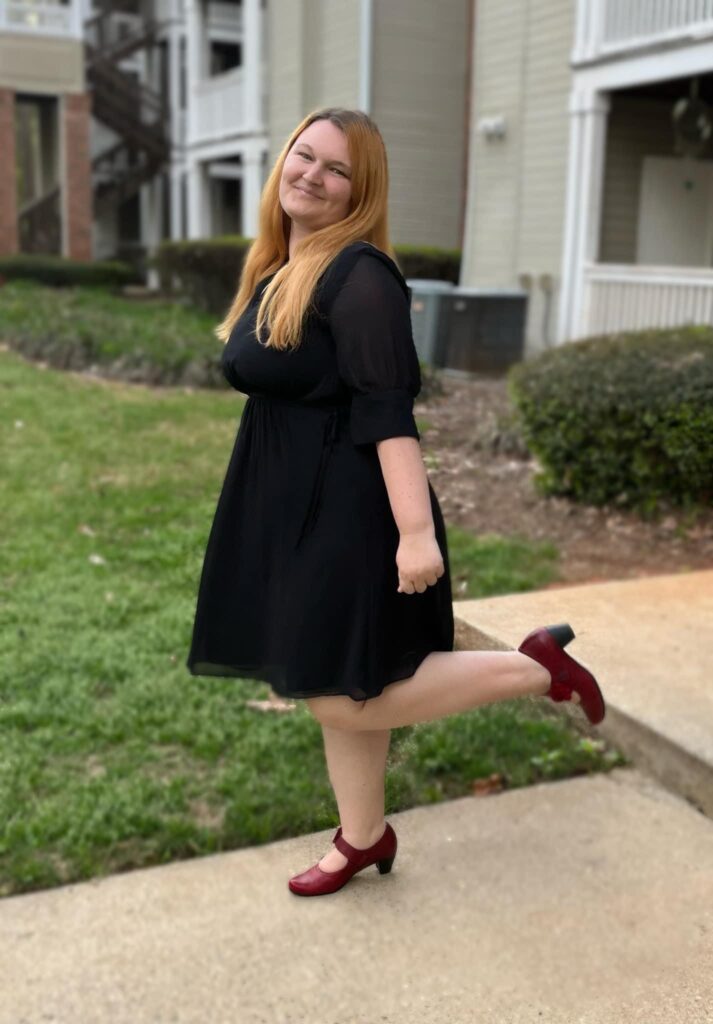 A woman with auburn hair wears a black dress and red heels and lifts one foot behind her.