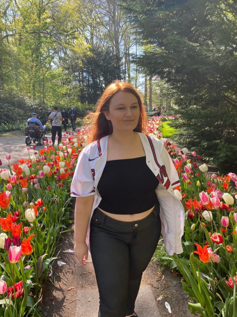 A woman with auburn hair, wearing an open Atlanta Braves jersey over a black tank top and jeans, walk on a sidewalk with rows of tulips on each side.