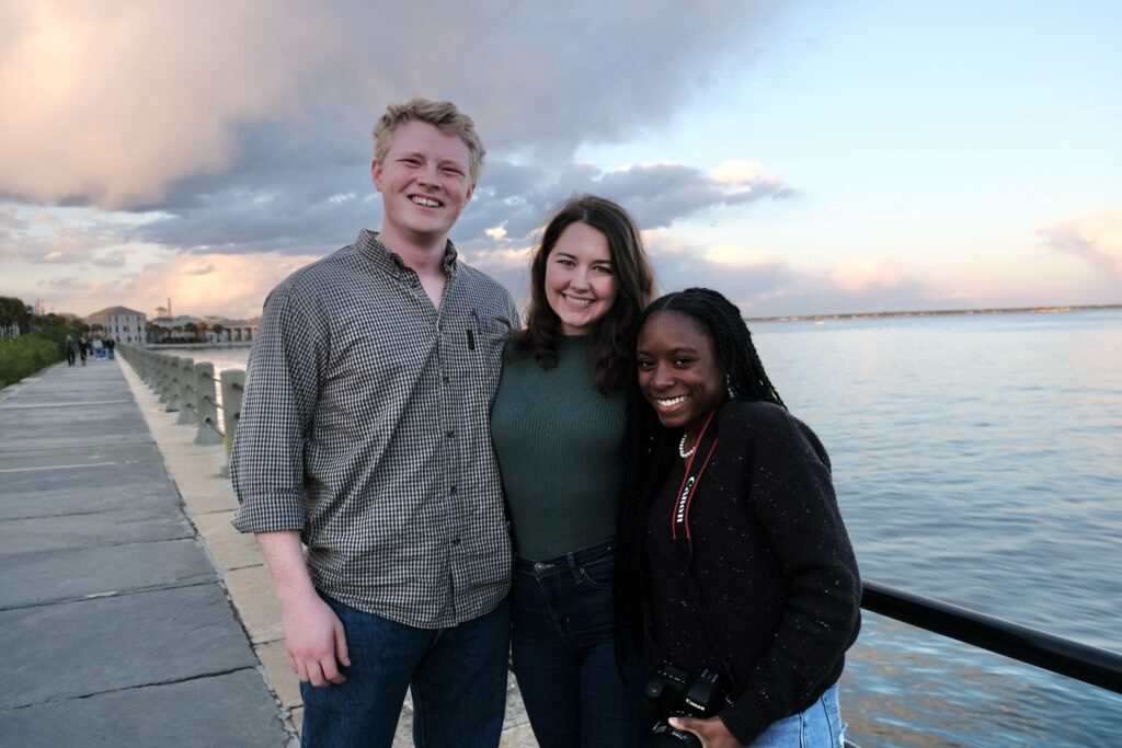 A male college student and two female college students pose for a photo, with the ocean in the background.